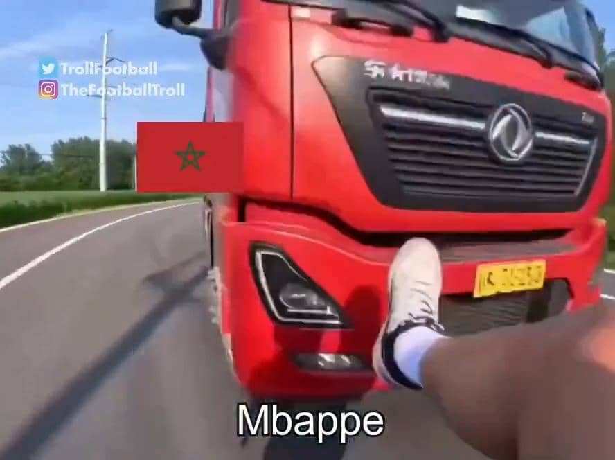 Truk merah dengan bendera Maroko di bagian depan melaju di jalan, sementara seseorang dengan tulisan Mbappe tampak di depannya.