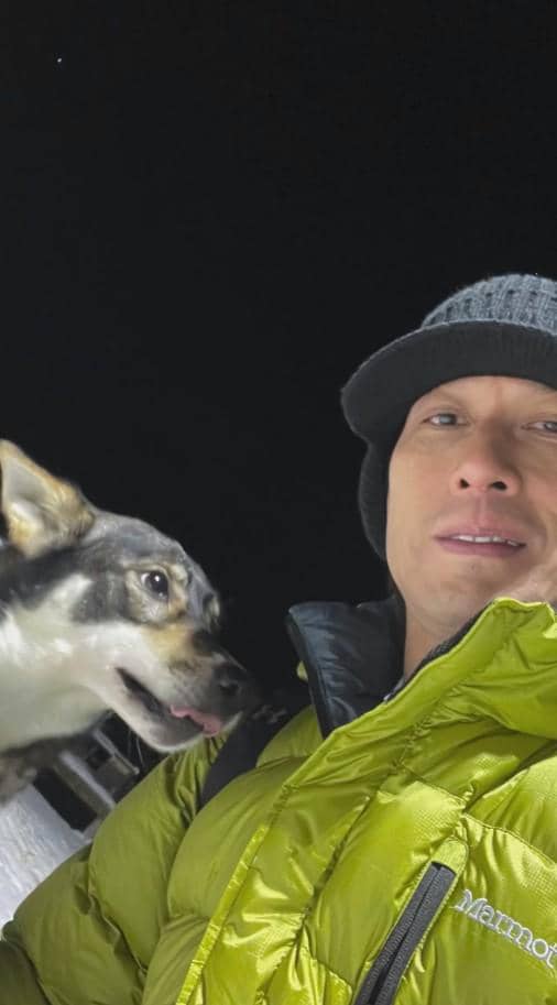 Seorang pria mengenakan jaket hijau dan topi hitam berpose di malam hari bersama seekor anjing di area bersalju di Norwegia.