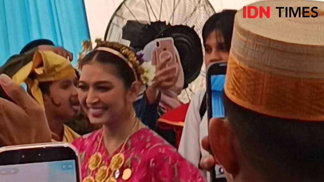 Seorang perempuan mengenakan busana tradisional berwarna merah muda dengan hiasan kepala bunga, tersenyum di tengah kerumunan orang yang mengambil foto.