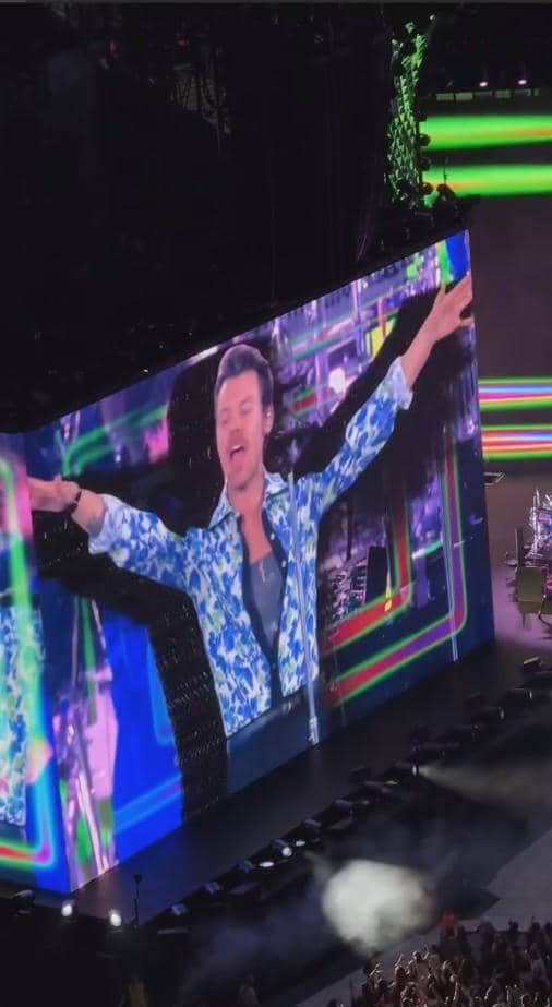 Layar besar menampilkan penyanyi pria mengenakan kemeja bermotif biru saat konser dengan pencahayaan warna-warni di panggung.