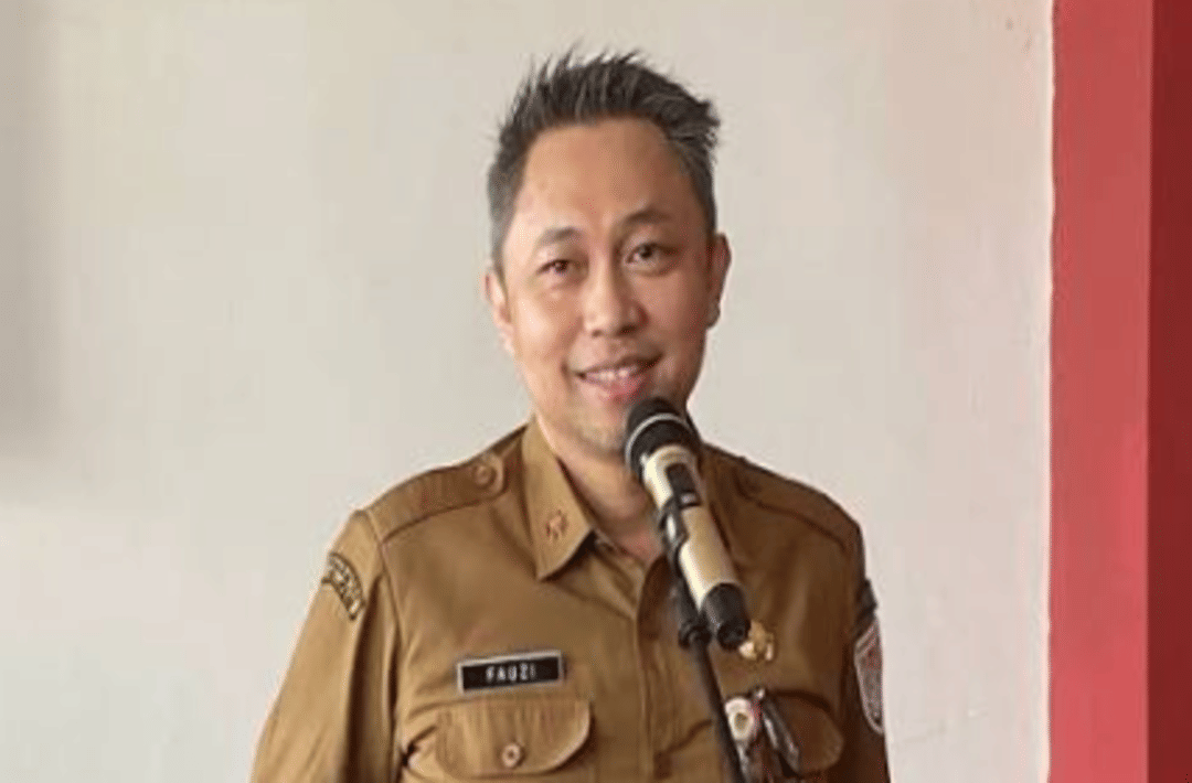 Seorang pria mengenakan seragam dinas cokelat sedang berbicara di depan mikrofon di dalam ruangan dengan latar dinding putih dan merah.