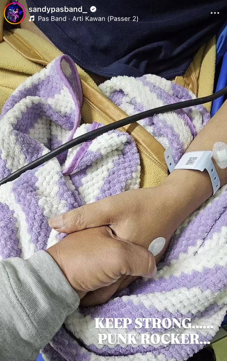 Dua tangan saling menggenggam di atas selimut ungu putih, salah satunya memakai gelang pasien rumah sakit dan kabel medis.