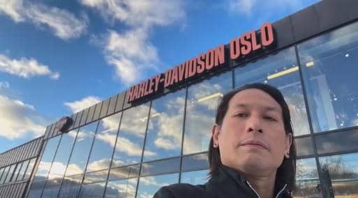 Seorang pria berpose di depan gedung bertuliskan Harley-Davidson Oslo dengan langit biru dan awan di latar belakang.