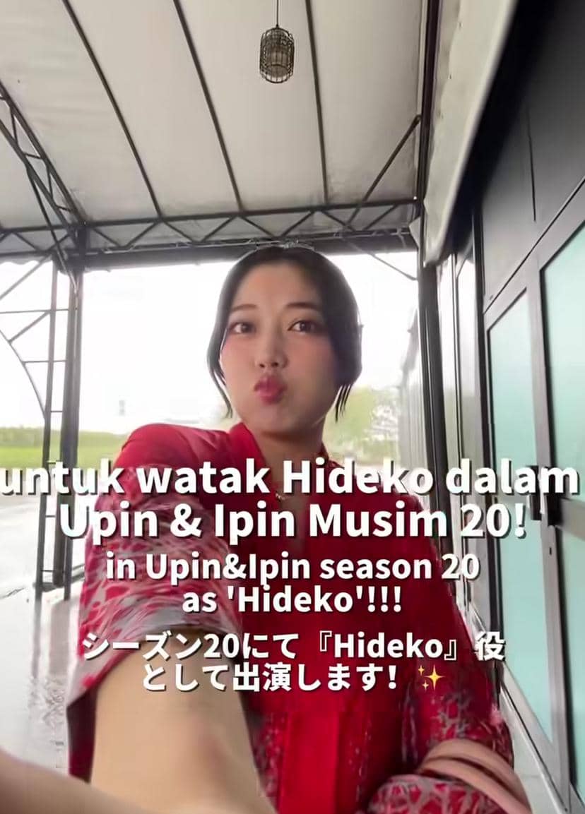 Seorang perempuan mengenakan pakaian merah berpose selfie di area beratap logam dengan teks pengumuman peran Hideko di Upin & Ipin Musim 20.
