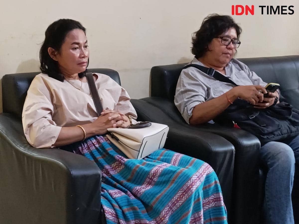 Dua perempuan duduk di sofa hitam, salah satunya mengenakan rok bermotif warna biru dan krem, sementara yang lain memegang ponsel.