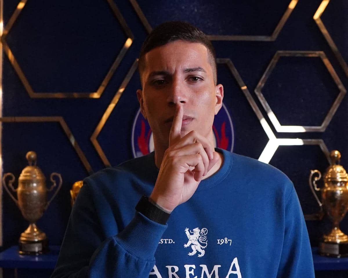 Seorang pria mengenakan sweatshirt biru bertuliskan Arema berdiri di depan latar trofi emas dengan jari di depan bibirnya.
