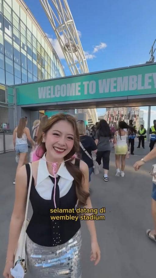 Seorang wanita tersenyum mengenakan pakaian bergaya modern di depan gerbang bertuliskan 'Welcome to Wembley' dengan suasana ramai pengunjung.