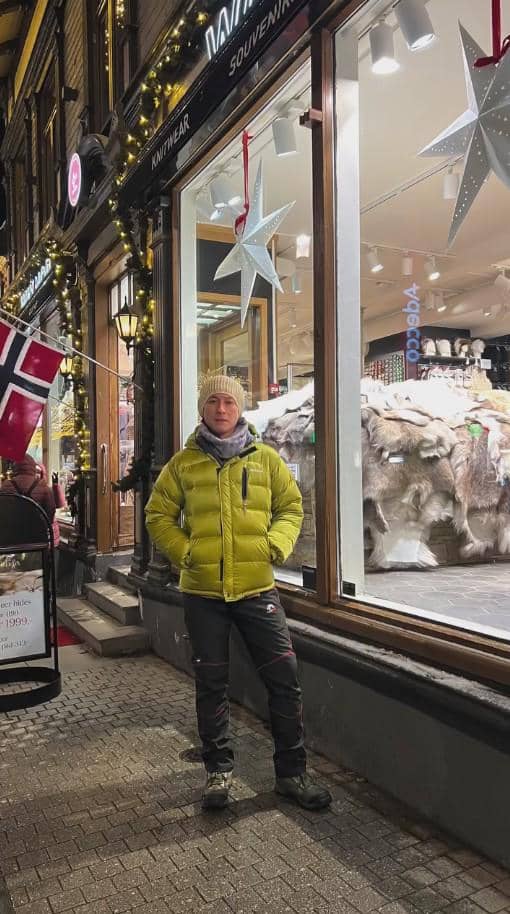 Seorang pria mengenakan jaket hijau dan topi rajut berdiri di trotoar depan toko suvenir berhias lampu dan bendera Norwegia.