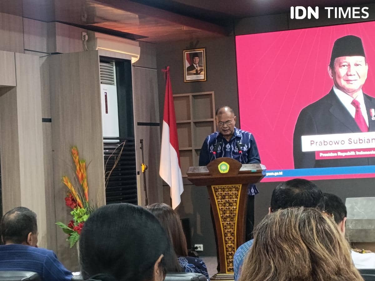 Seorang pejabat berbicara di podium dalam ruangan dengan layar besar menampilkan gambar tokoh nasional dan bendera Indonesia di sampingnya.