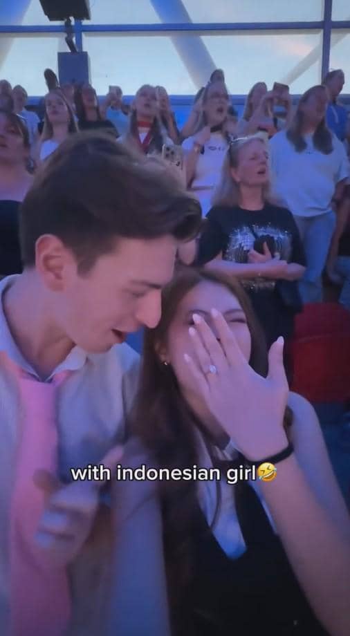 Seorang pria dan wanita tertawa bersama di tengah kerumunan penonton konser, dengan suasana meriah dan pencahayaan biru di sekitar mereka.