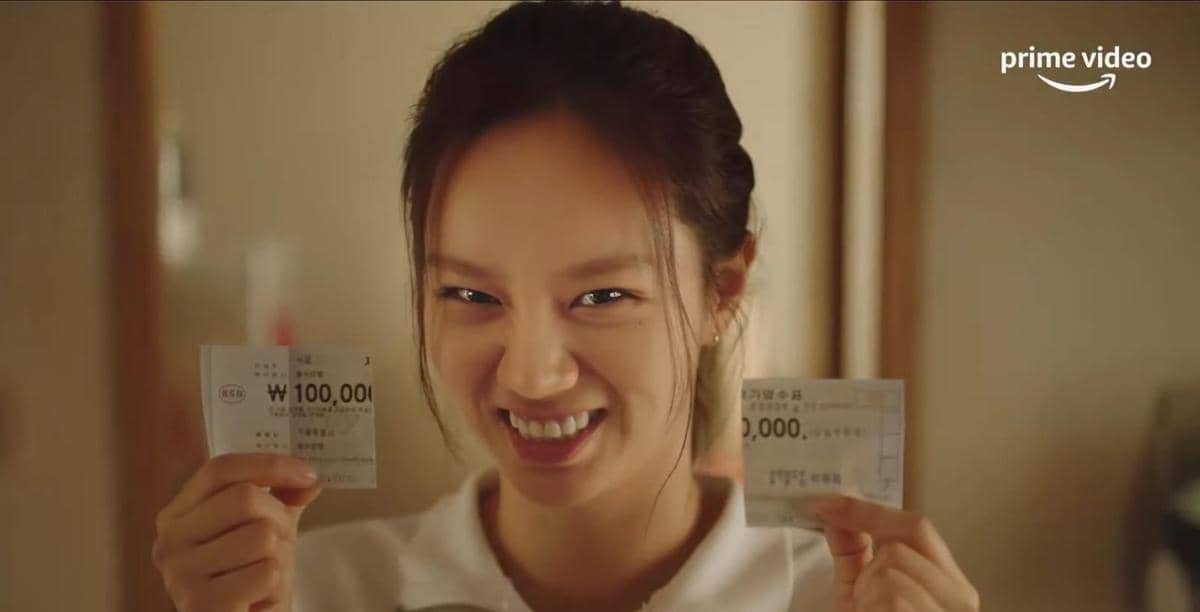 Seorang wanita tersenyum sambil memegang dua lembar uang kertas Korea di tangannya dalam cuplikan drama May I Help You.