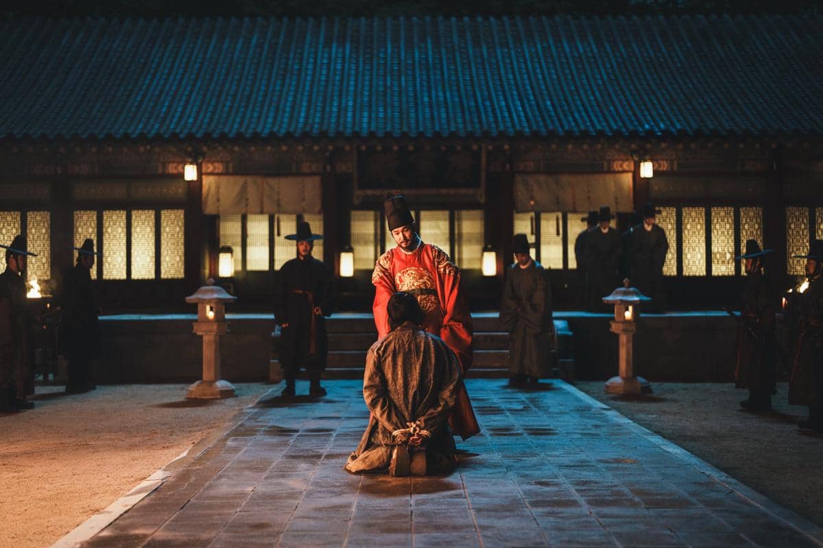 Seorang pria berpakaian kerajaan merah berdiri di depan seseorang yang berlutut di halaman istana pada malam hari dalam adegan drama The East Palace.