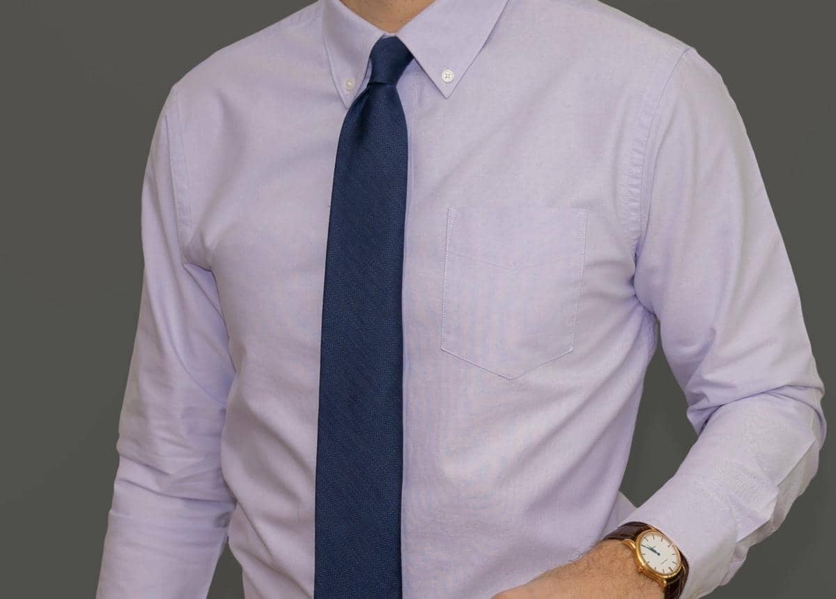 Pria mengenakan kemeja Oxford berwarna ungu muda dengan dasi biru tua dan jam tangan cokelat di pergelangan tangan kiri.