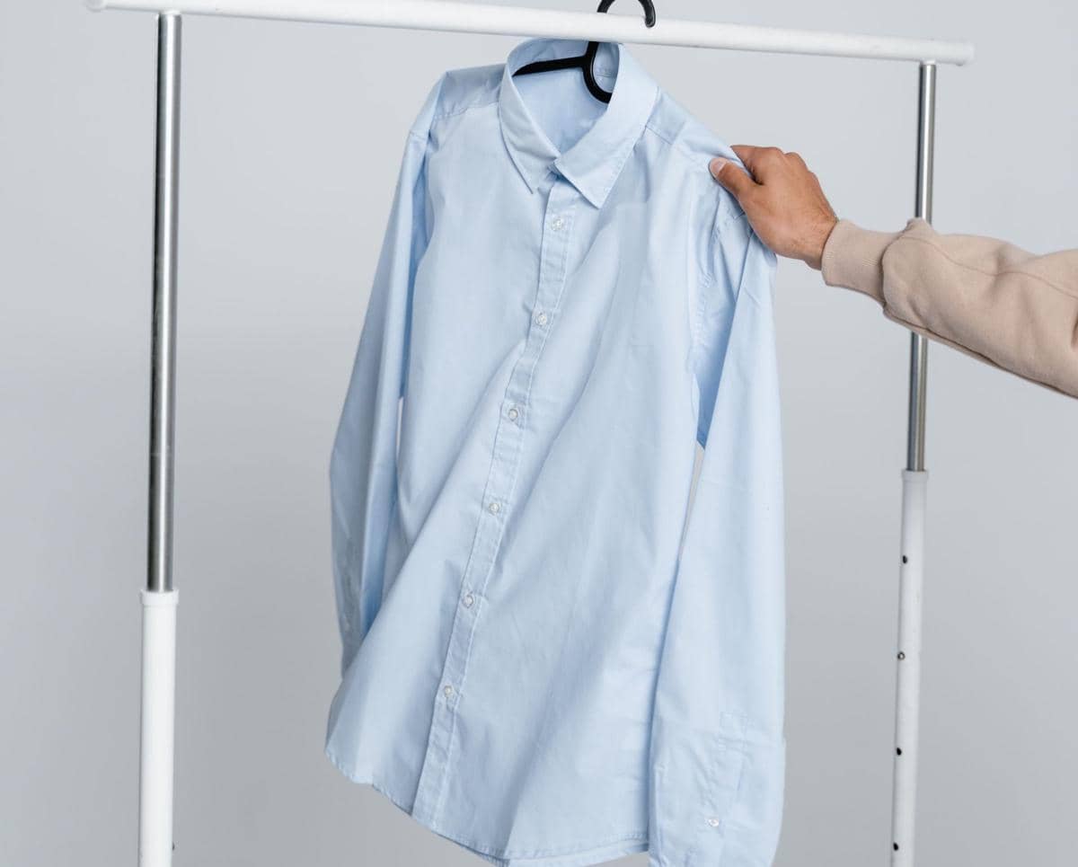 Seseorang memegang kemeja Oxford berwarna biru muda yang tergantung di gantungan pakaian dengan latar belakang abu-abu polos.