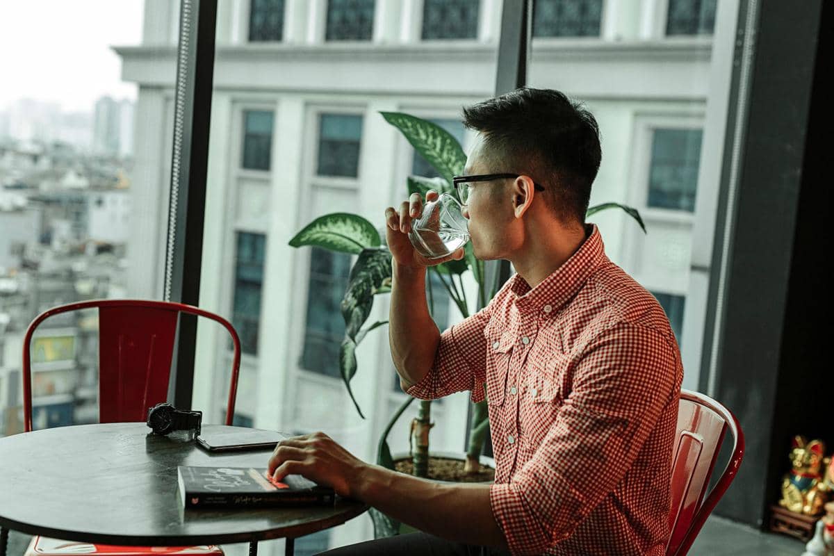 Pria mengenakan kemeja Oxford kotak-kotak merah duduk di kafe dekat jendela besar sambil memegang gelas minuman.