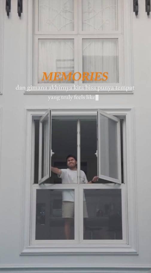 Seorang pria berdiri di jendela rumah berwarna putih dengan desain klasik, mengenakan kaus putih dan celana pendek.
