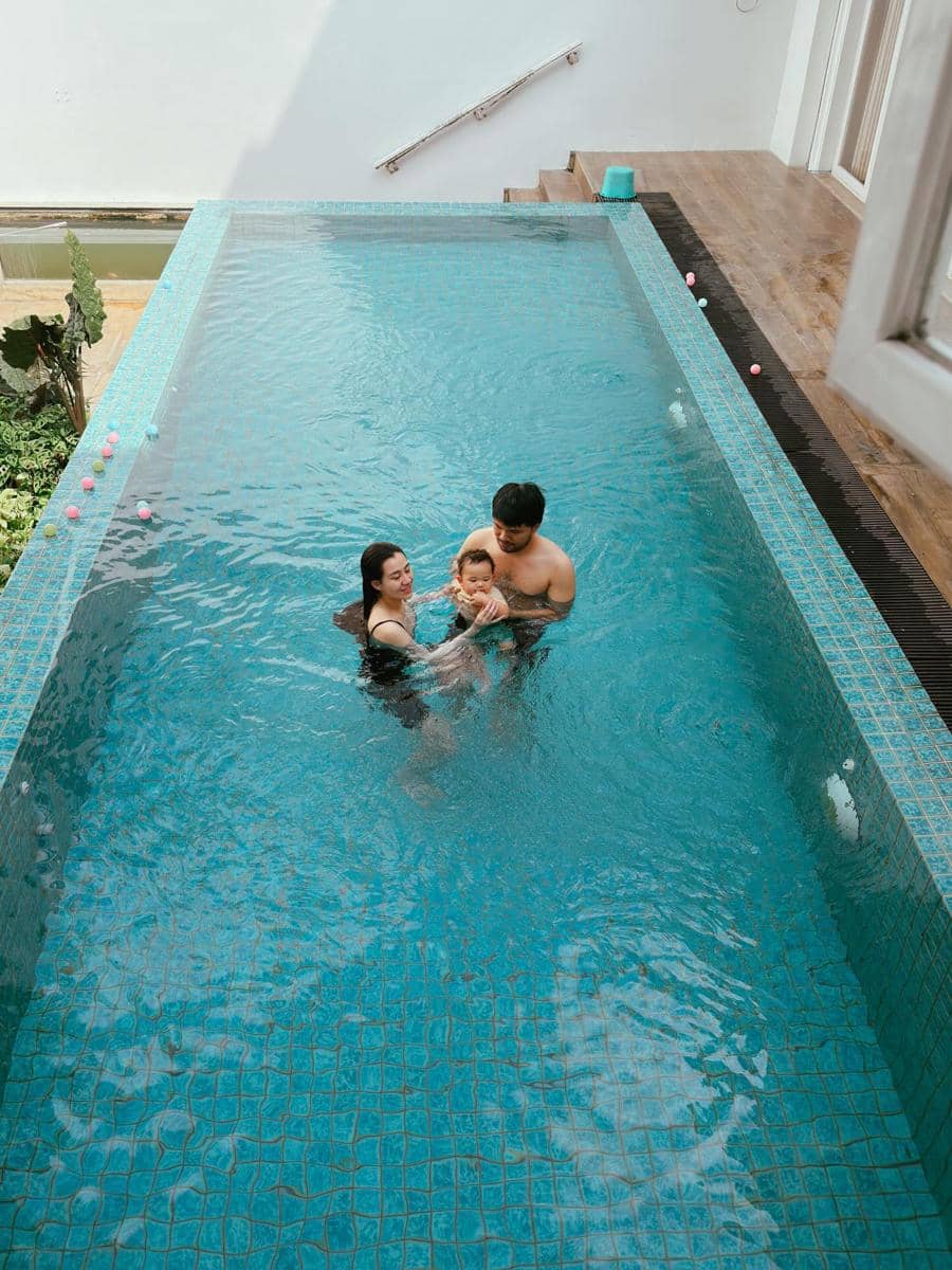 Sepasang orang dewasa bermain air bersama seorang bayi di kolam renang rumah dengan dinding putih dan lantai kayu di tepi kolam.