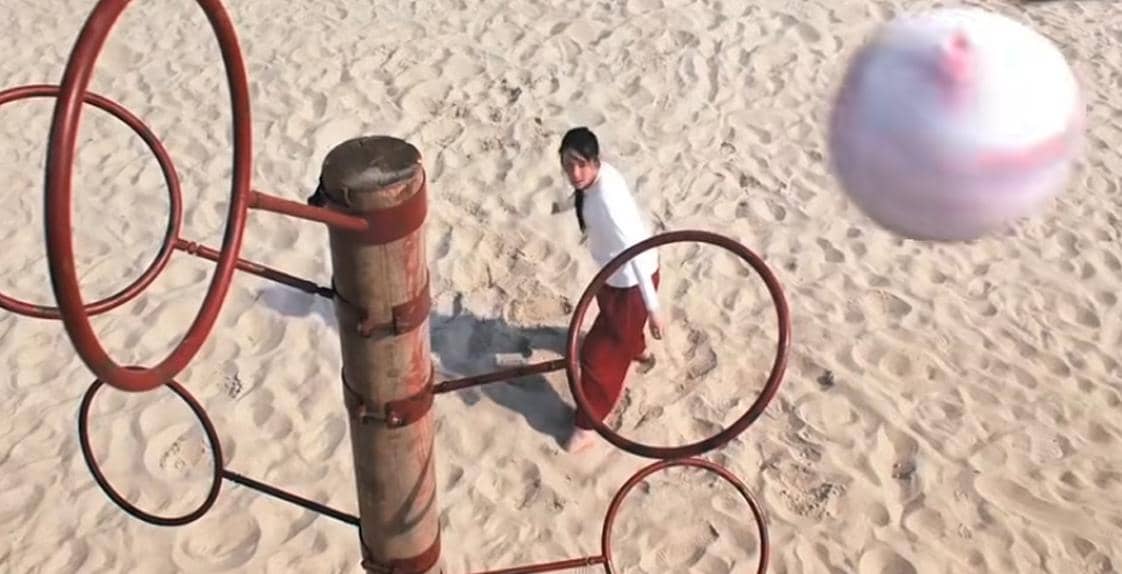 Seorang pemain mengenakan pakaian putih dan merah berlatih kungfu sambil bermain bola di atas pasir dengan alat latihan berbentuk lingkaran logam.