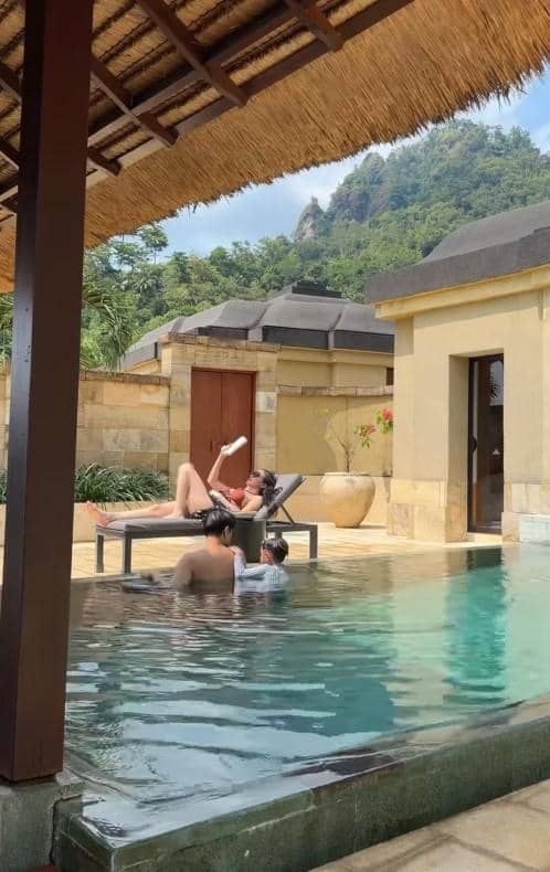 Seorang wanita berjemur di kursi tepi kolam renang sementara dua orang bermain air di vila bergaya tropis dengan latar perbukitan hijau.