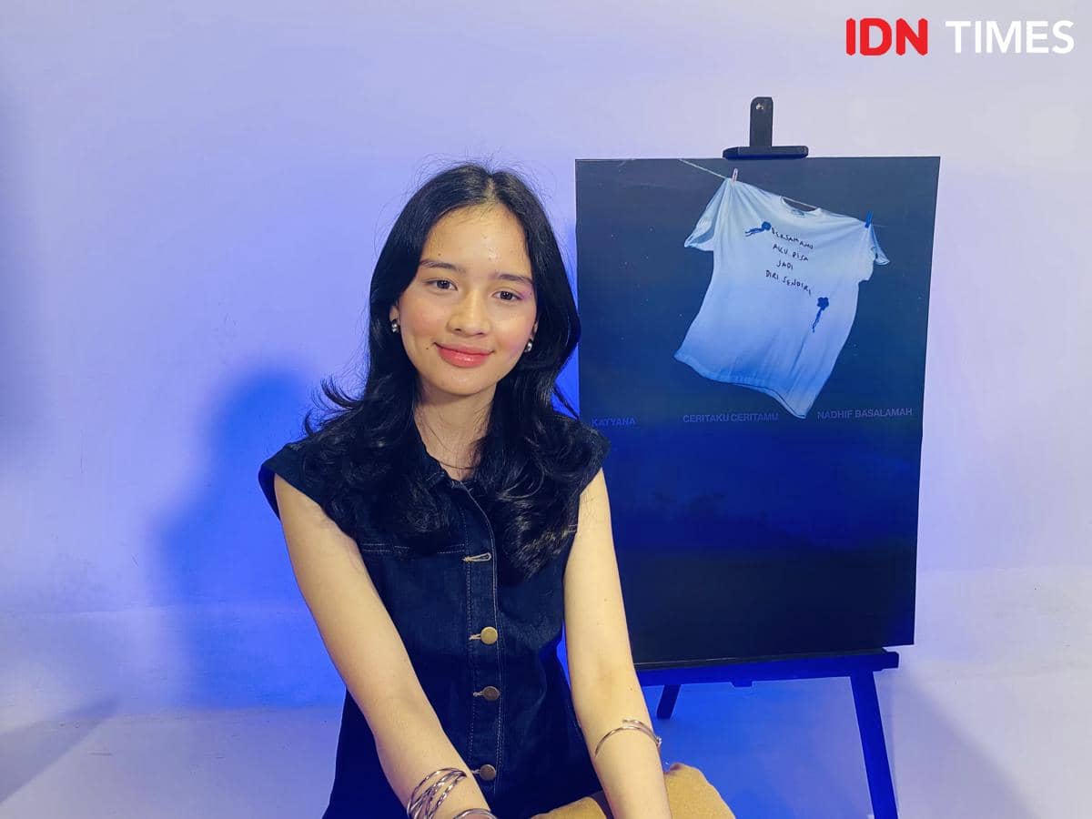 Seorang perempuan berambut panjang duduk tersenyum di depan poster bertema biru dengan gambar kaus putih bertulisan tangan di atasnya.