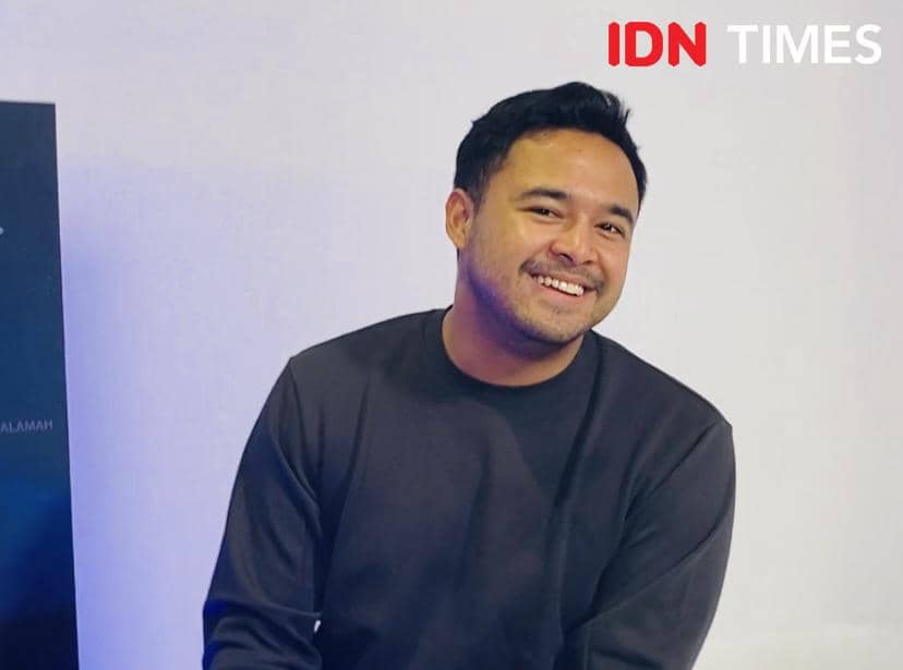 Seorang pria mengenakan sweater hitam tersenyum sambil duduk di kantor Trinity Optima Production dengan latar dinding putih.