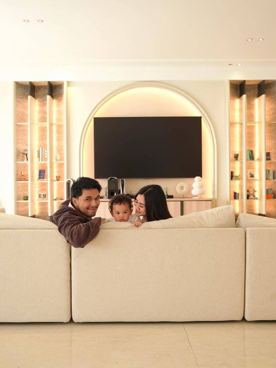 Seorang pria, wanita, dan anak kecil bersandar di sofa krem di ruang tamu modern dengan pencahayaan hangat dan televisi besar di dinding.