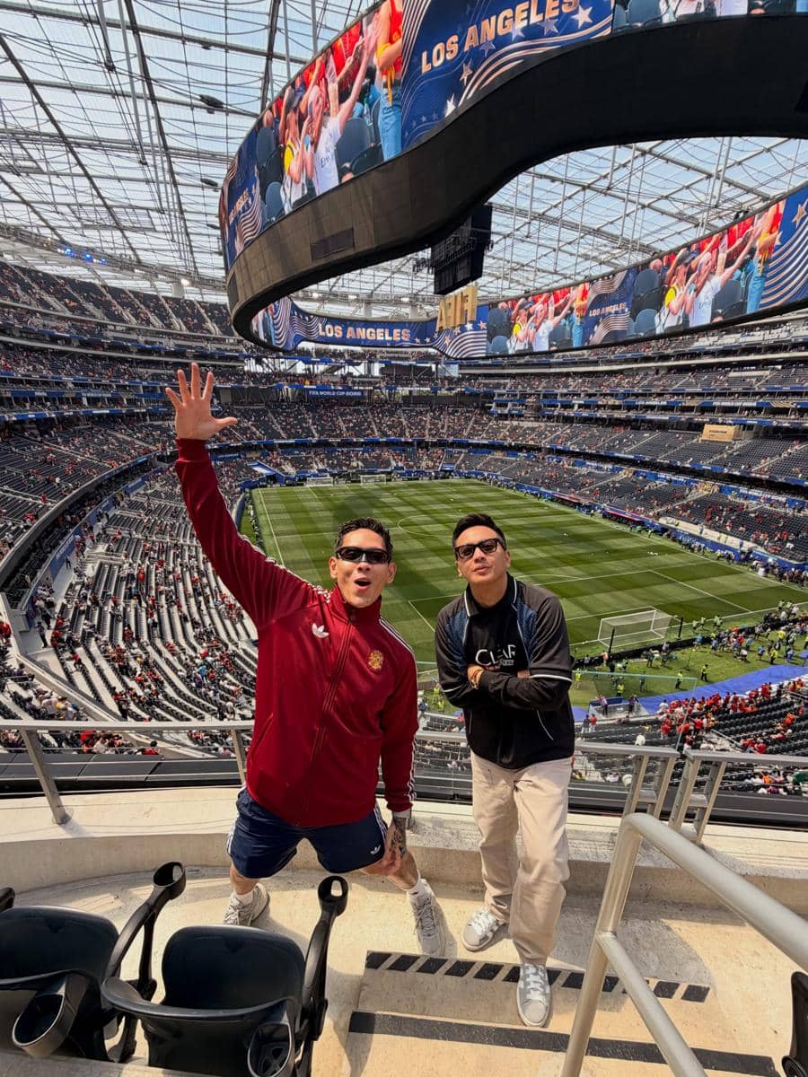 Dua pria berpose di tribun stadion besar di Los Angeles dengan latar lapangan sepak bola menjelang pertandingan Spanyol vs Belgia.