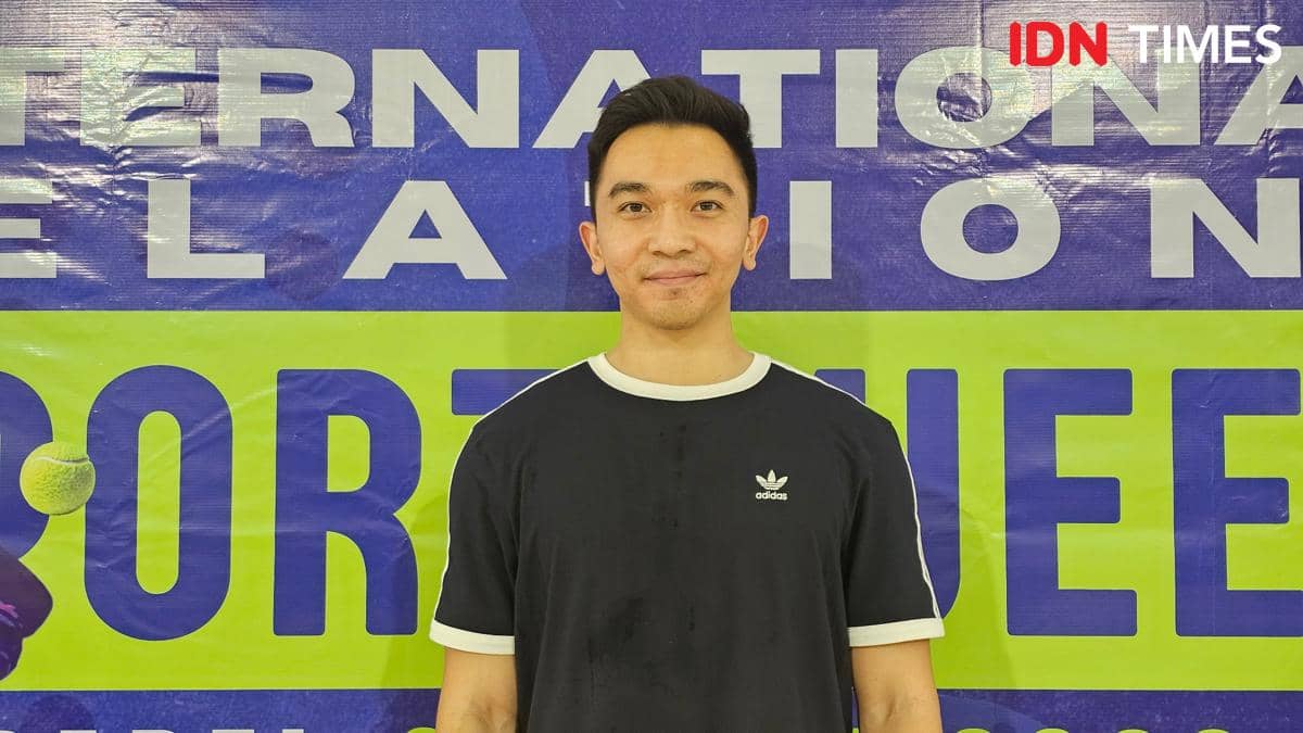 Seorang pria mengenakan kaus hitam dengan logo Adidas berdiri di depan latar bertuliskan INTERNATIONAL RELATION SPORT WEEK berwarna biru dan hijau.