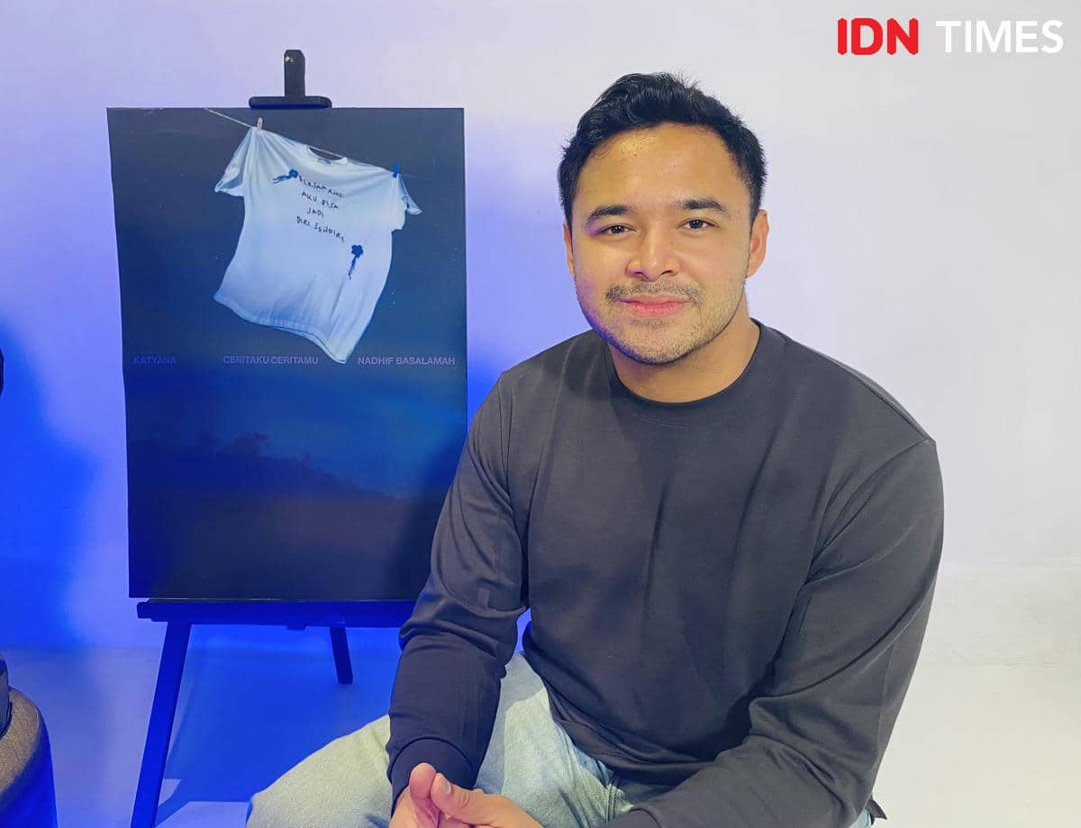Seorang pria duduk tersenyum di depan poster perilisan lagu berjudul 'Ceritaku Ceritamu' dalam konferensi pers di kantor Trinity Optima Production.