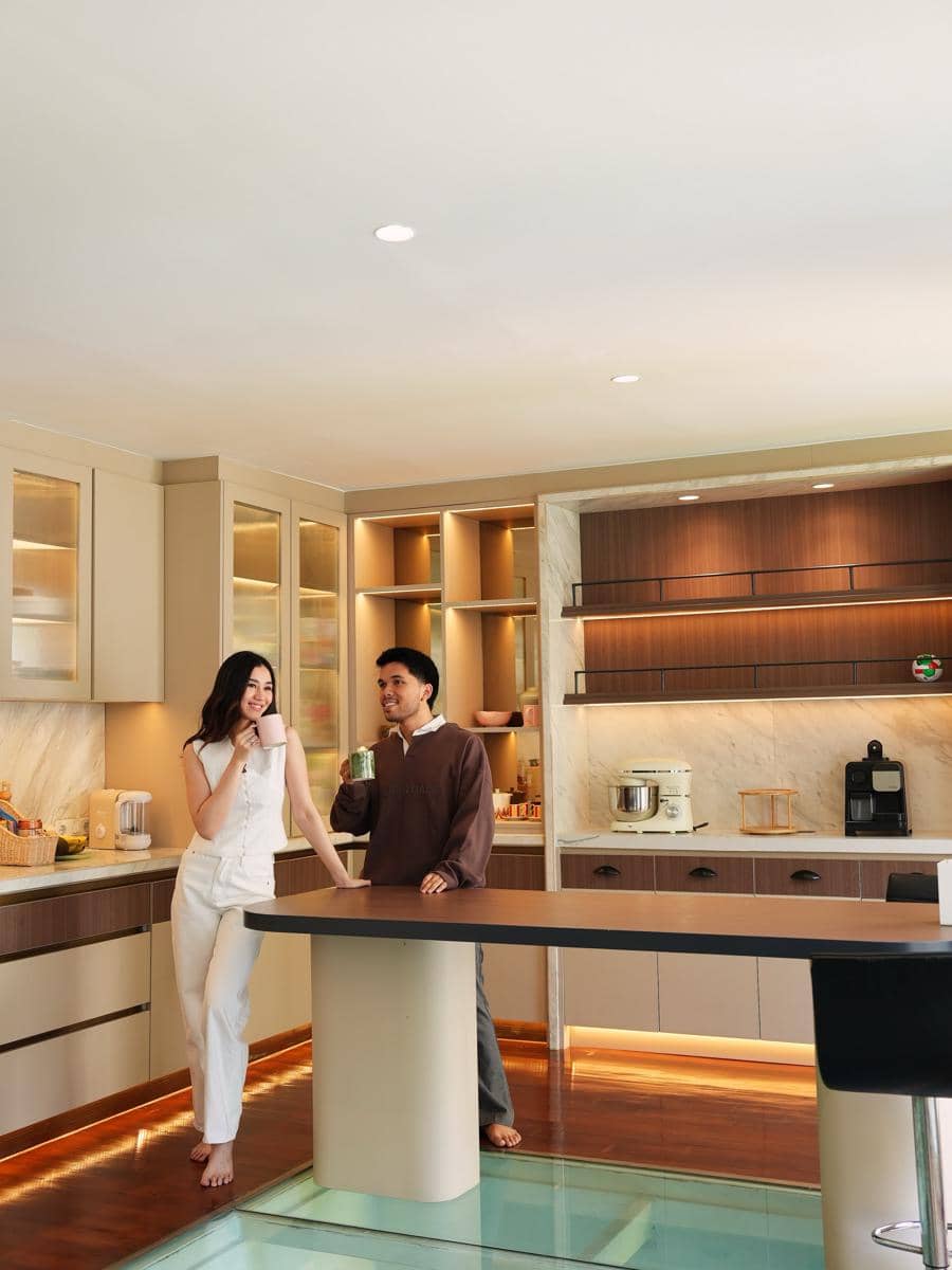 Dua orang berdiri santai di dapur modern dengan pencahayaan hangat, meja tengah besar, dan lantai kaca transparan.