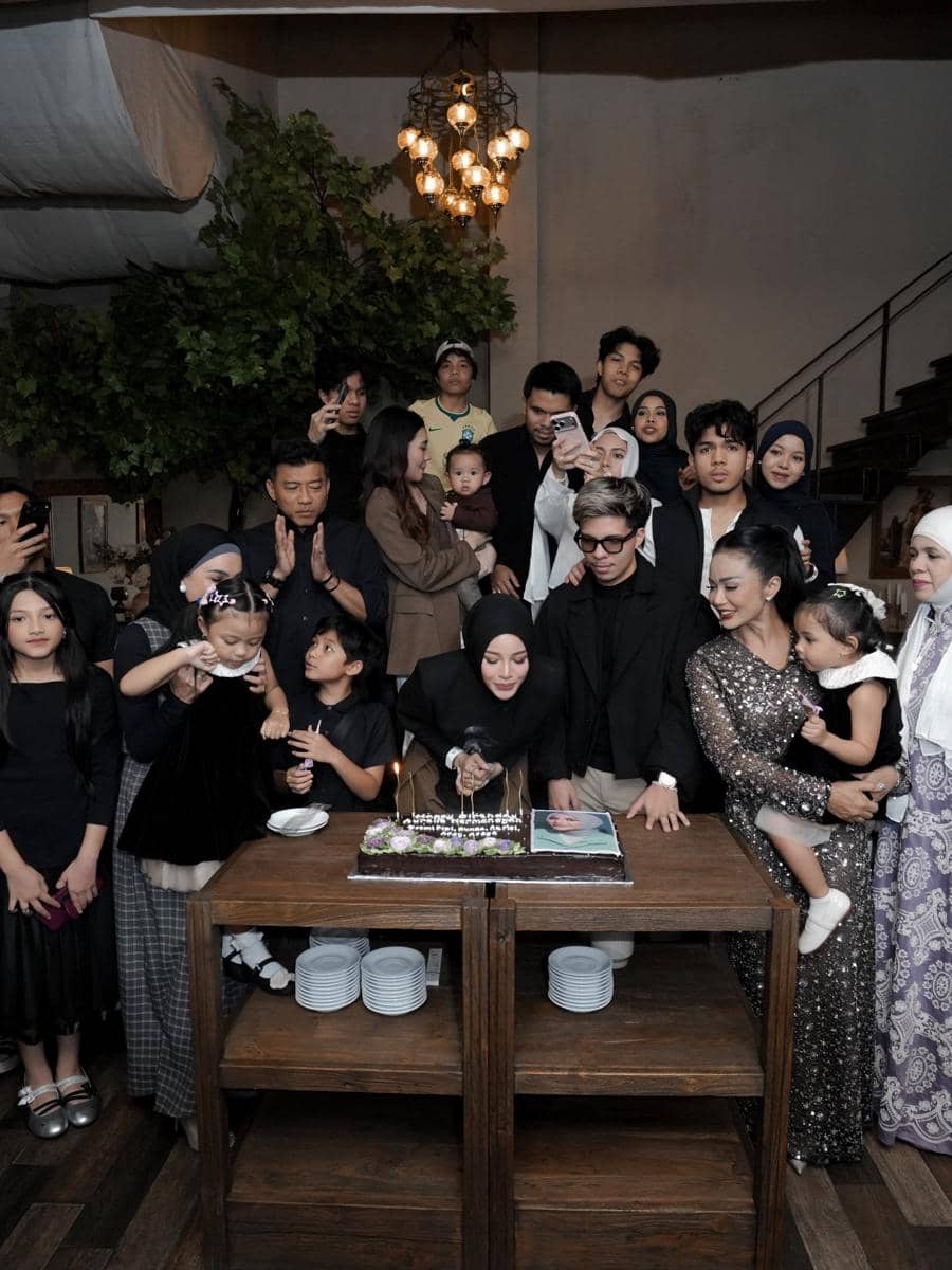 Sekelompok orang berkumpul di sekitar meja dengan kue ulang tahun, beberapa anak kecil ikut merayakan di ruangan bernuansa hangat.
