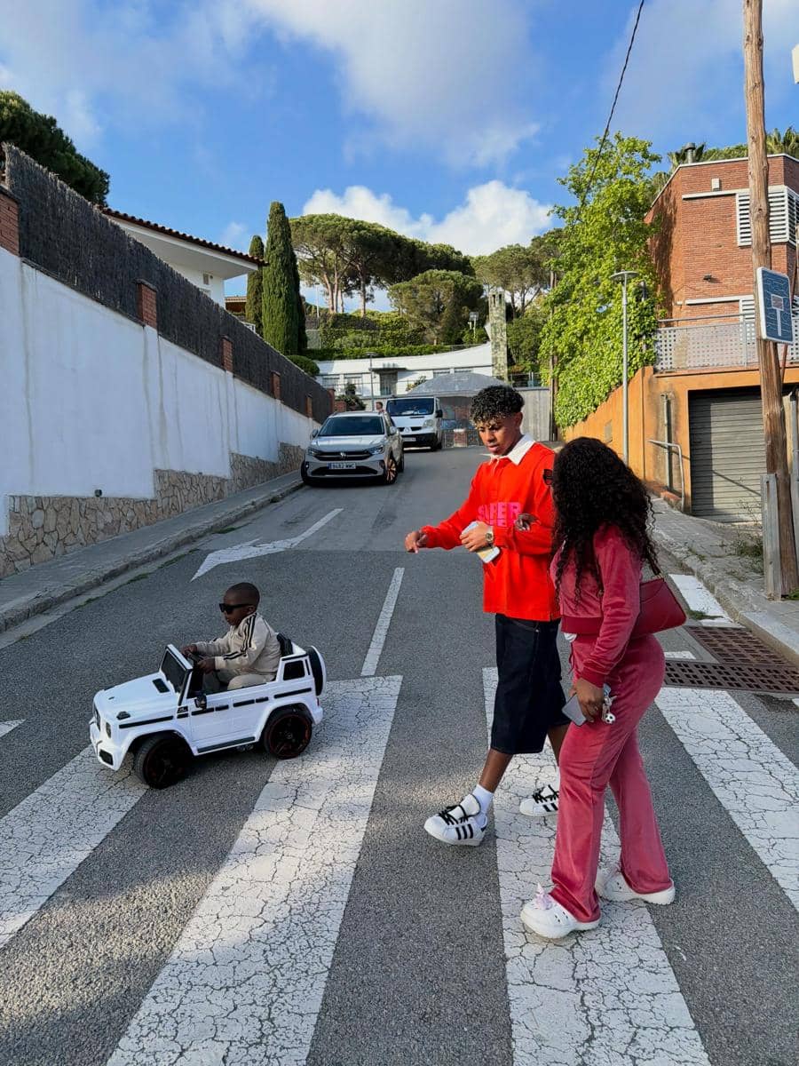 Seorang anak kecil mengendarai mobil mainan putih di jalan perumahan, sementara dua orang dewasa berdiri di dekatnya di penyeberangan jalan.