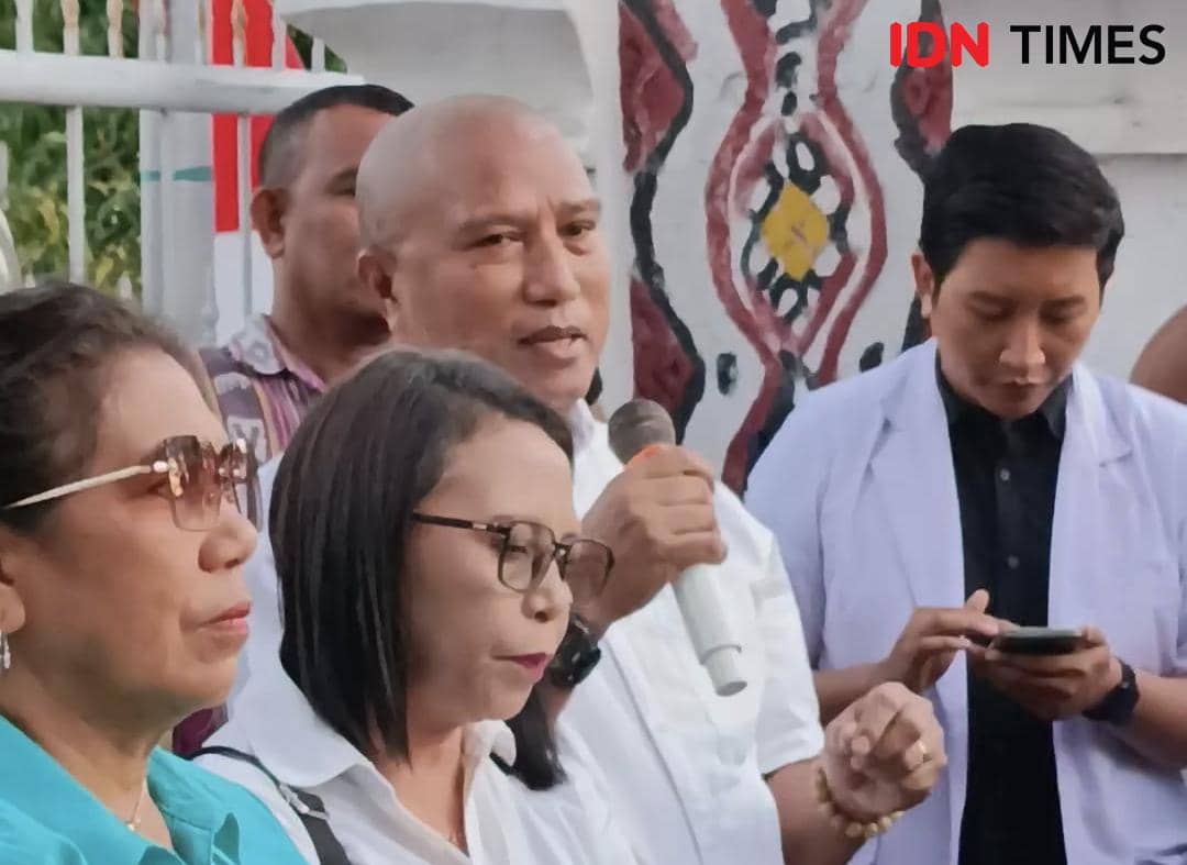 Seorang pria memegang mikrofon berbicara di depan beberapa orang, dengan latar belakang dinding bermotif tradisional dan pagar putih.