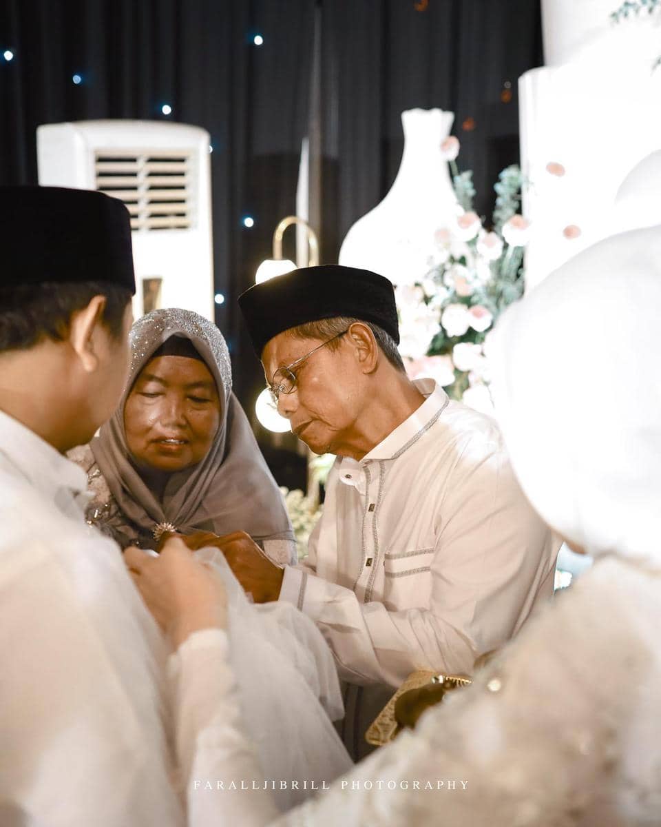 Seorang pria lanjut usia mengenakan baju putih dan peci hitam bersama seorang wanita berhijab abu-abu dalam acara pernikahan.