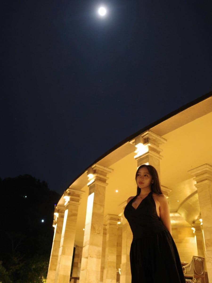 Seorang wanita mengenakan gaun hitam berdiri di depan bangunan berpilar dengan pencahayaan hangat di bawah sinar bulan penuh.