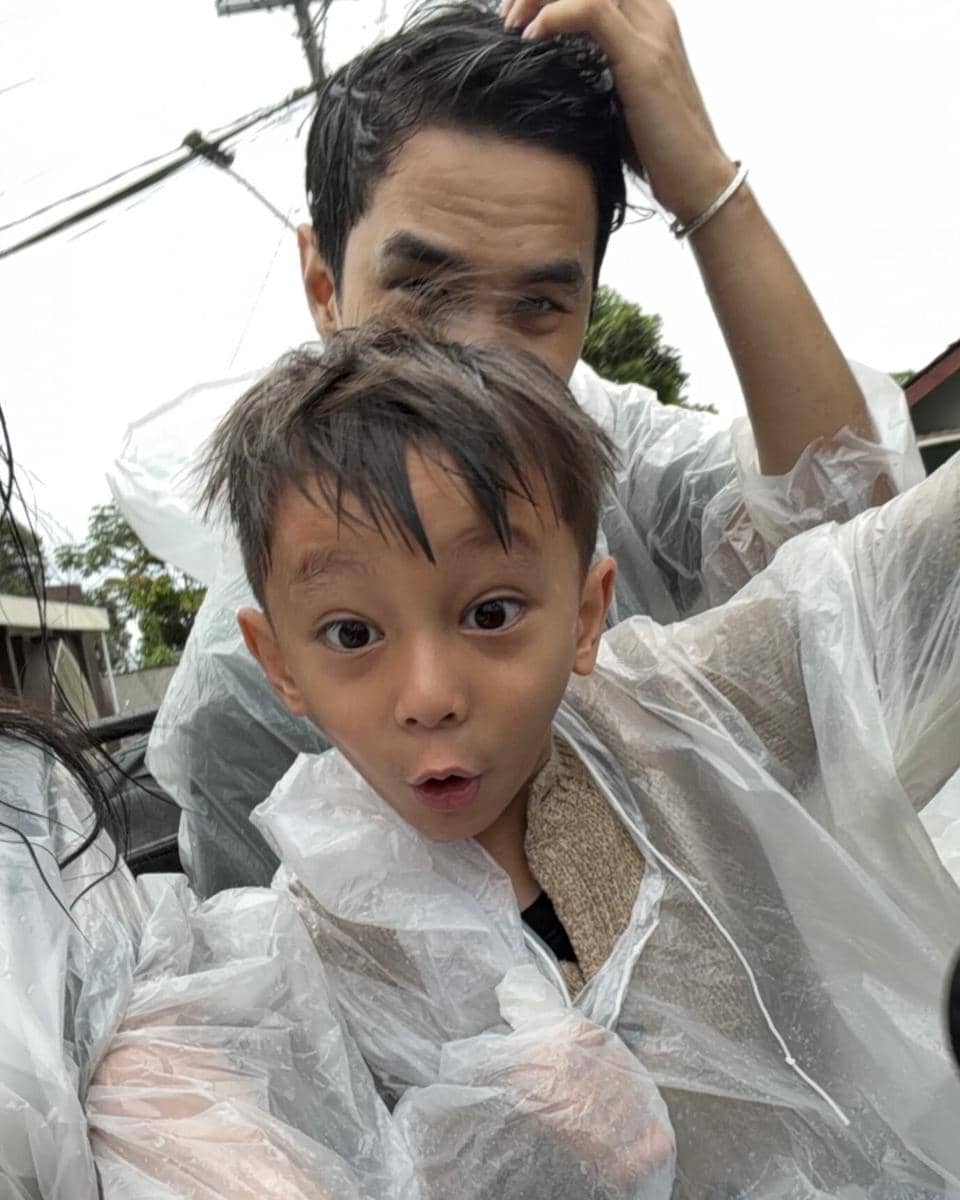 Seorang anak dan pria mengenakan jas hujan plastik transparan tampak tersenyum dan berpose ceria di luar ruangan saat cuaca basah.
