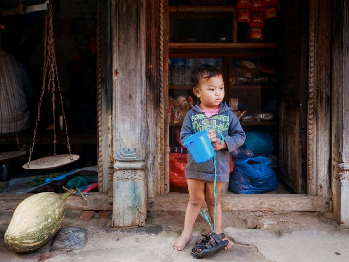 Seorang anak kecil berdiri di depan pintu rumah kayu sambil memegang ember biru, mengenakan sweter abu-abu dan tanpa alas kaki.