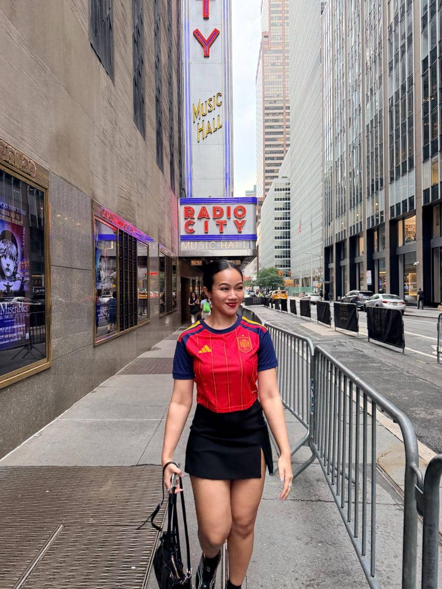 Seorang perempuan mengenakan jersey timnas Spanyol berjalan di trotoar dekat Radio City Music Hall di antara gedung-gedung tinggi kota.