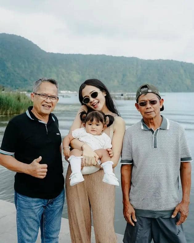 Seorang wanita memegang bayi sambil berpose bersama dua pria di tepi danau dengan latar pegunungan hijau.