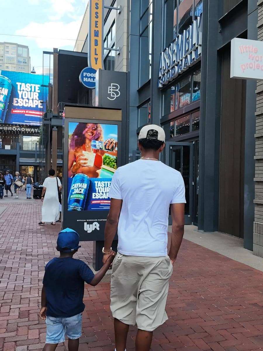 Seorang pria dan anak kecil berjalan bergandengan tangan di area pejalan kaki kota dengan papan reklame dan gedung bertuliskan Assembly Food Hall.
