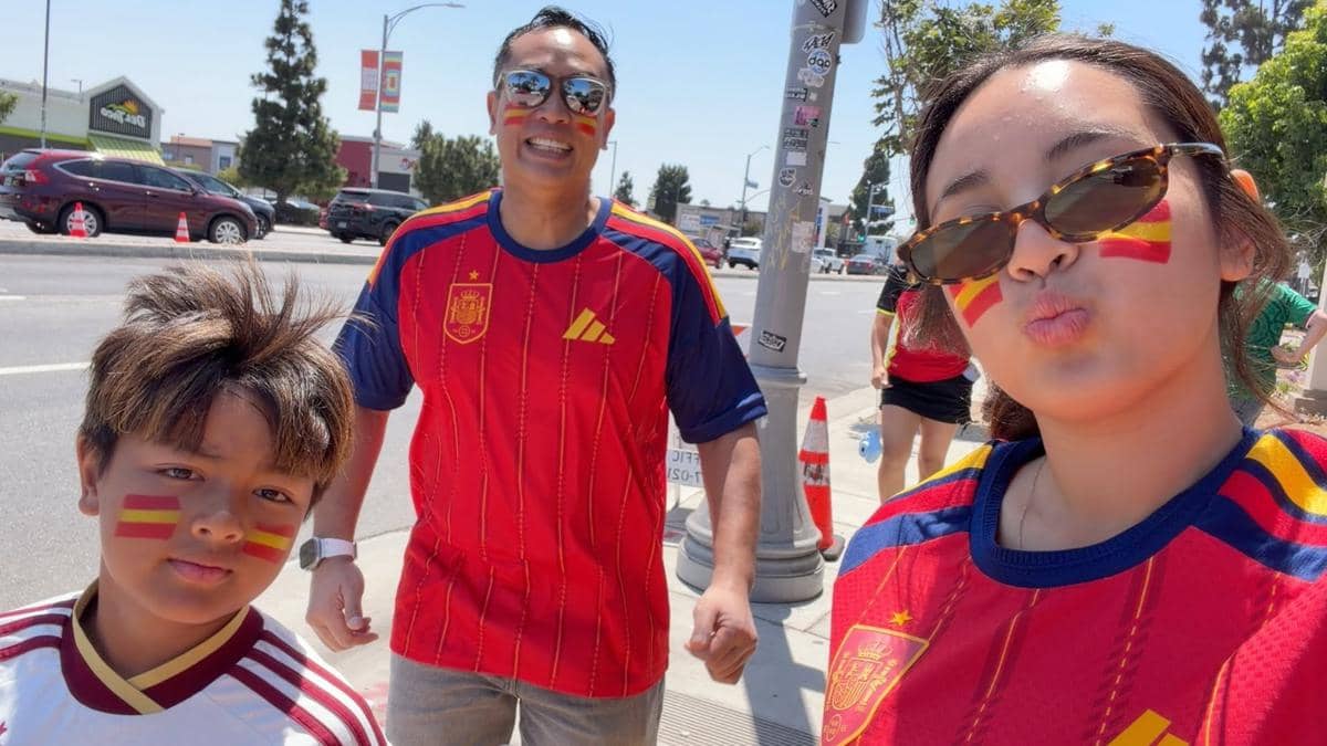 Tiga orang mengenakan jersey timnas Spanyol dengan wajah dicat bendera Spanyol, berpose di luar ruangan di bawah sinar matahari.