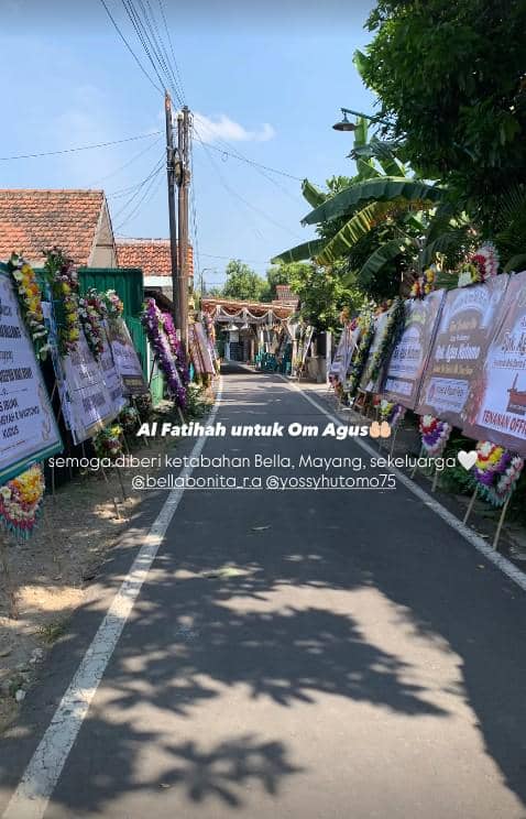 Jalan desa dipenuhi karangan bunga duka cita di kedua sisi, suasana pemakaman dengan langit cerah dan pepohonan di sekitar area.