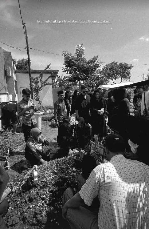 Sejumlah orang berpakaian hitam berkumpul di area pemakaman, beberapa duduk dan berdoa di dekat makam yang dipenuhi bunga.