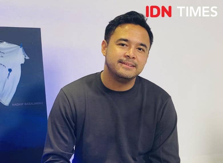 Seorang pria mengenakan sweater abu-abu gelap duduk di dalam ruangan dengan latar dinding putih dan poster berwarna biru di sampingnya.