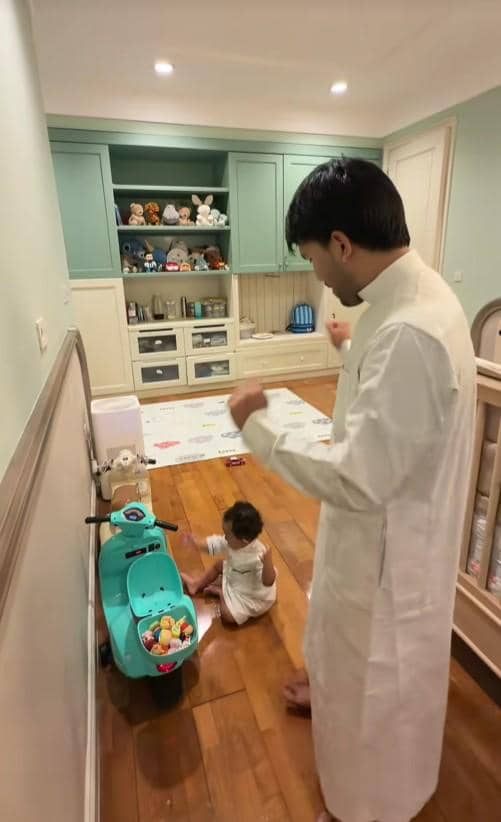 Seorang pria mengenakan pakaian putih berdiri di kamar anak dengan lantai kayu, sementara seorang balita duduk bermain di dekat mainan motor kecil berwarna hijau.