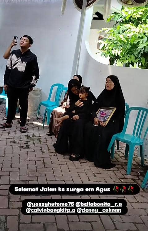 Beberapa orang berpakaian hitam duduk di kursi plastik biru saat prosesi pemakaman, seorang wanita memegang foto almarhum.