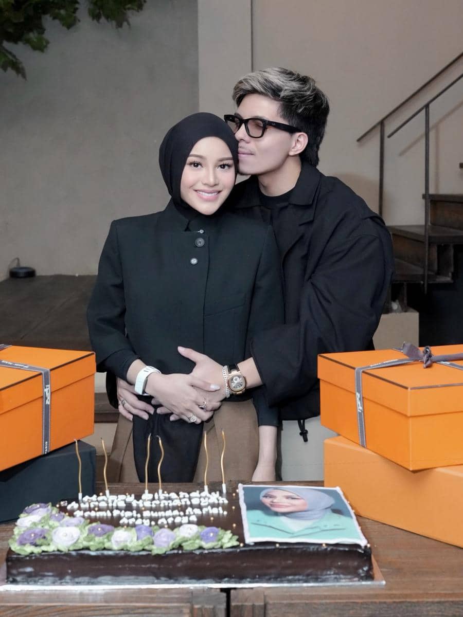 Seorang wanita berhijab dan pria berambut pirang berpose bersama di depan kue ulang tahun dan beberapa kotak hadiah berwarna oranye.