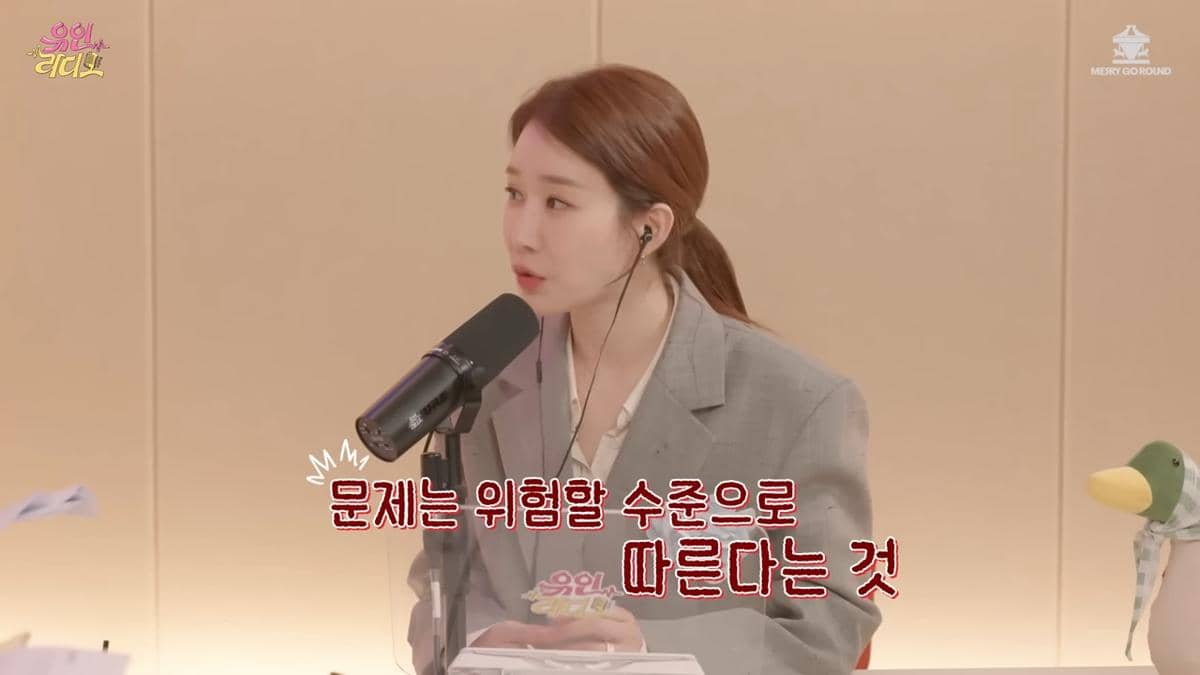 Seorang pembawa acara berbicara di depan mikrofon saat merekam episode podcast Yoo In Radio di studio dengan latar warna krem.