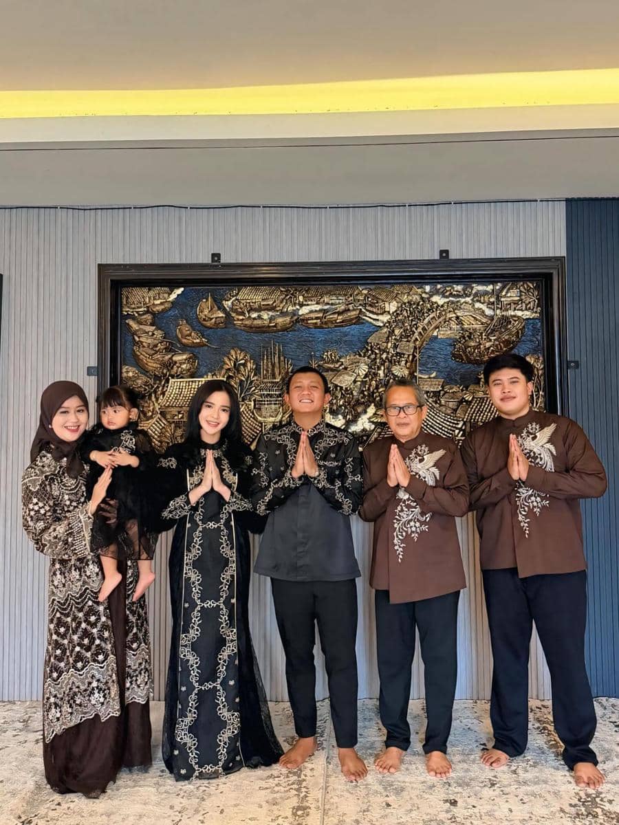 Enam anggota keluarga berpakaian senada warna cokelat dan hitam berpose sambil merapatkan tangan di depan ukiran dinding besar.
