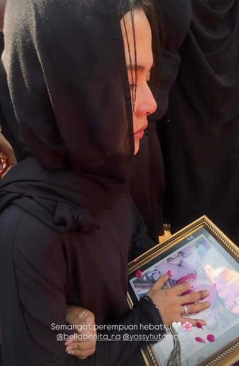 Seorang perempuan berpakaian hitam memegang bingkai foto dengan bunga mawar di atasnya saat menghadiri pemakaman di bawah sinar matahari.